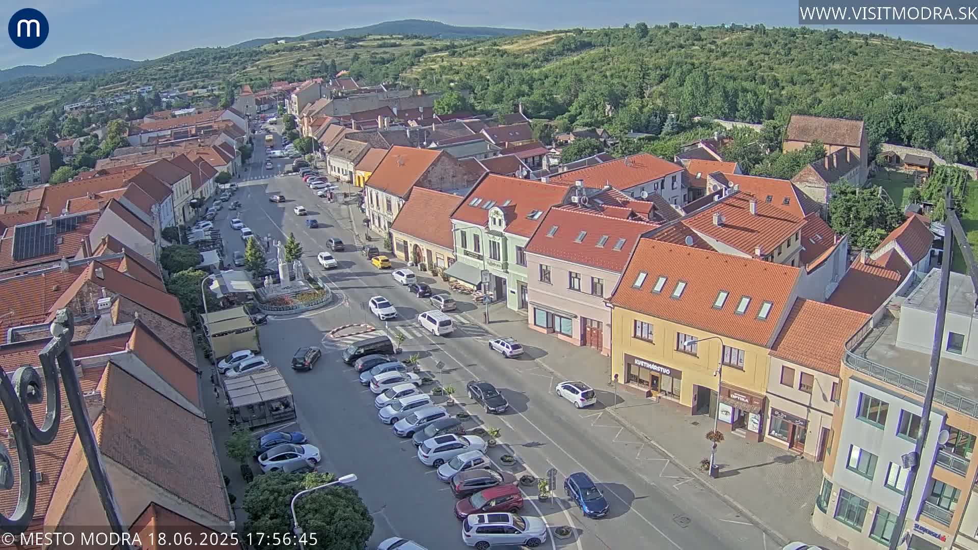 An aerial view of a town square and street lined with buildings on a sunny day, with cars parked along the side of the road and several vehicles driving through the town square.