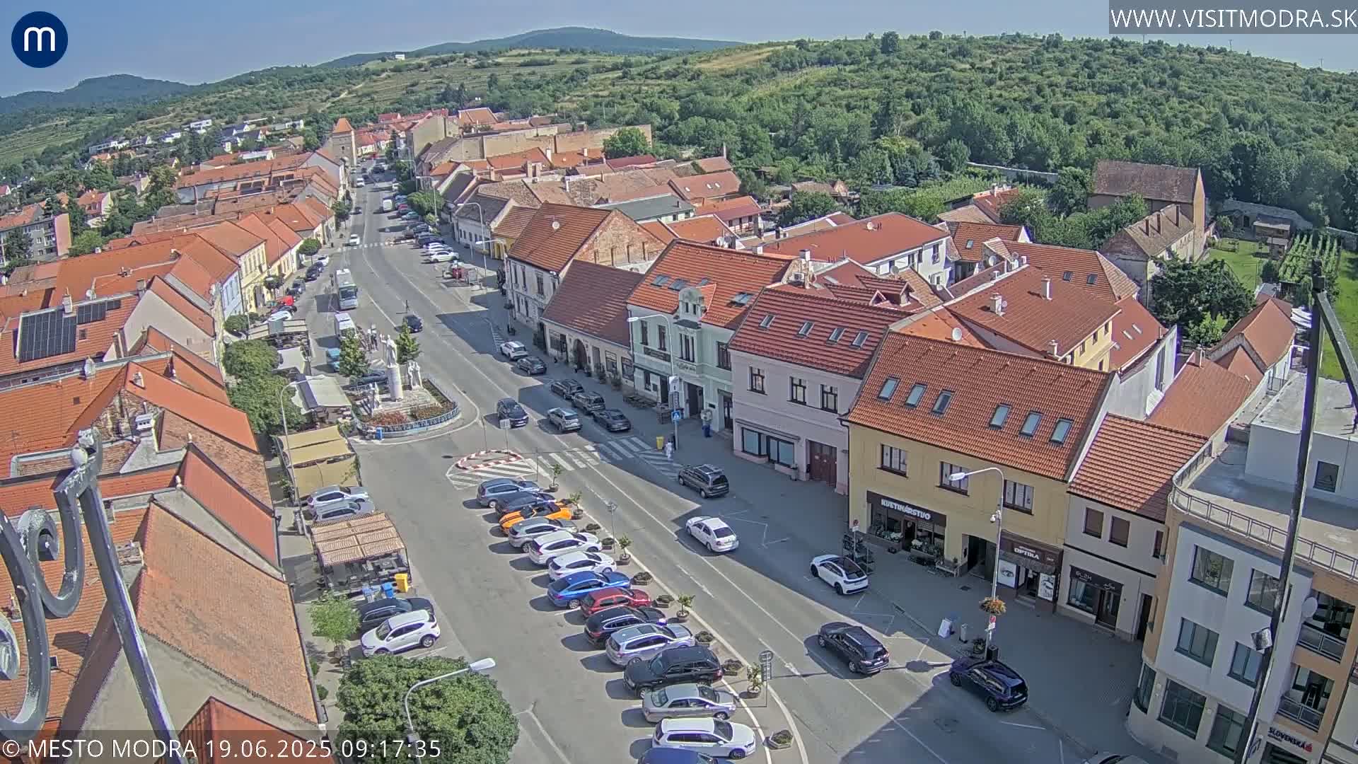 A sunny aerial view of a town's main street lined with parked cars and low-rise buildings,  surrounded by lush green hills.