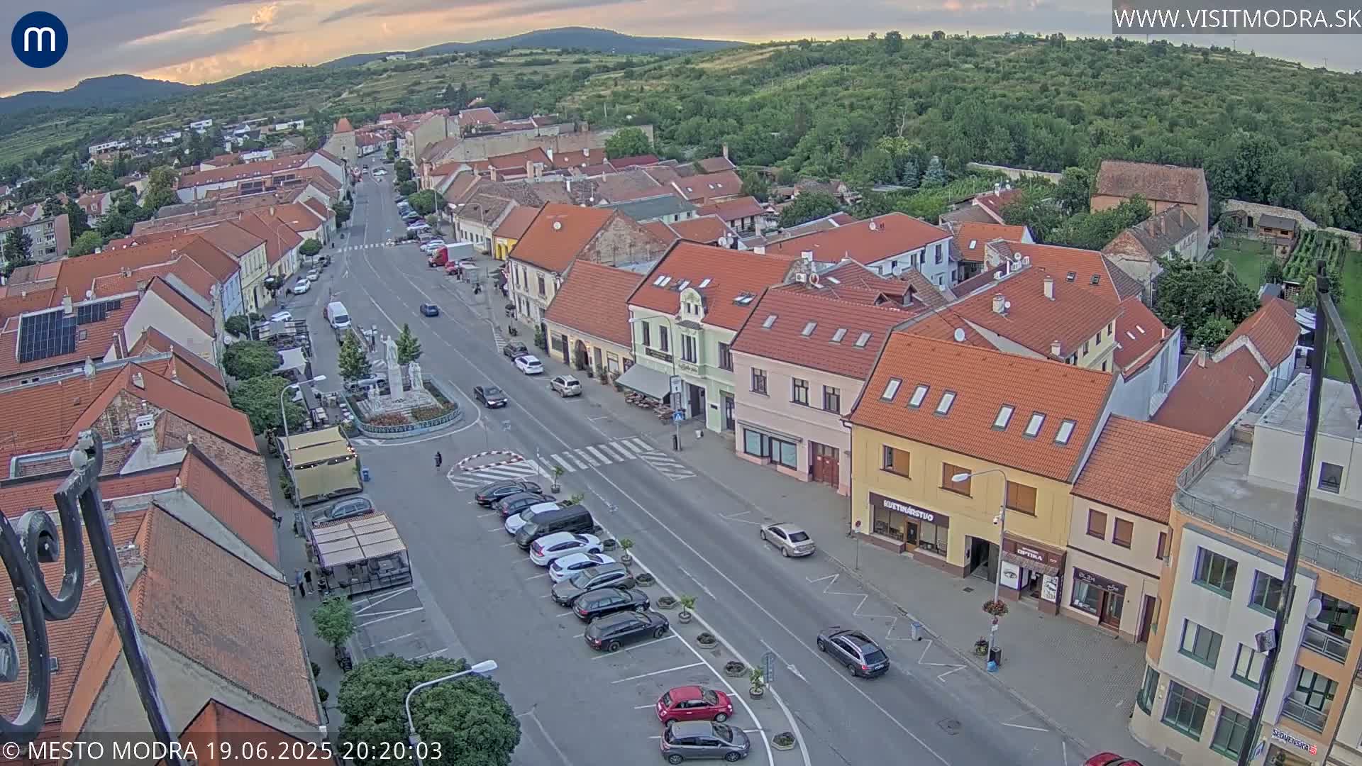 An aerial view of a town square with parked cars and buildings under a partly cloudy evening sky.