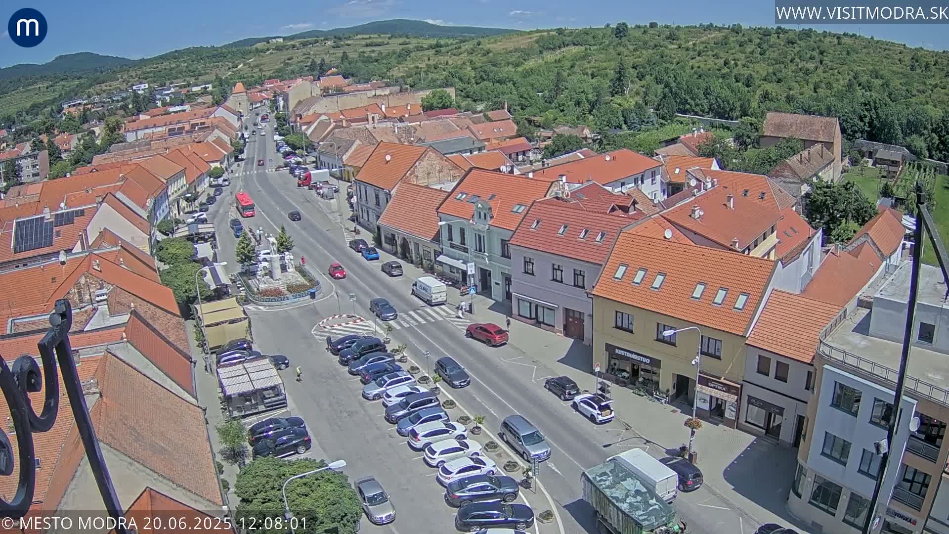 An aerial view of a town square with cars parked along the street, surrounded by buildings with red tile roofs under a sunny sky.