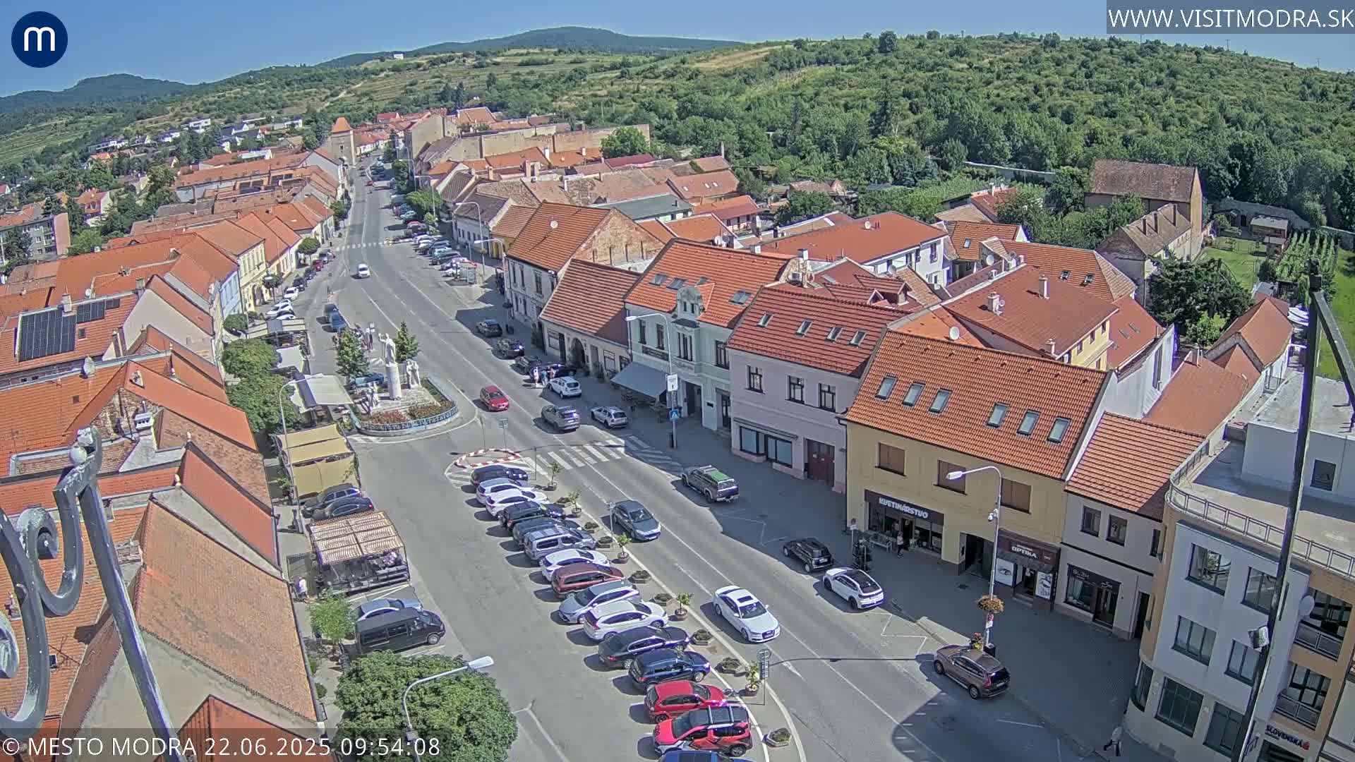 An aerial view of a town square on a sunny day, showing a street lined with parked cars and buildings with red-tiled roofs.