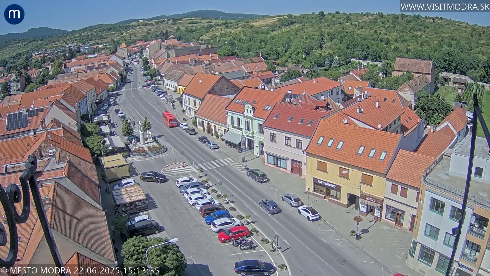 An aerial view shows a town square with parked cars, a street with cars driving, and buildings with red tile roofs under a sunny sky.