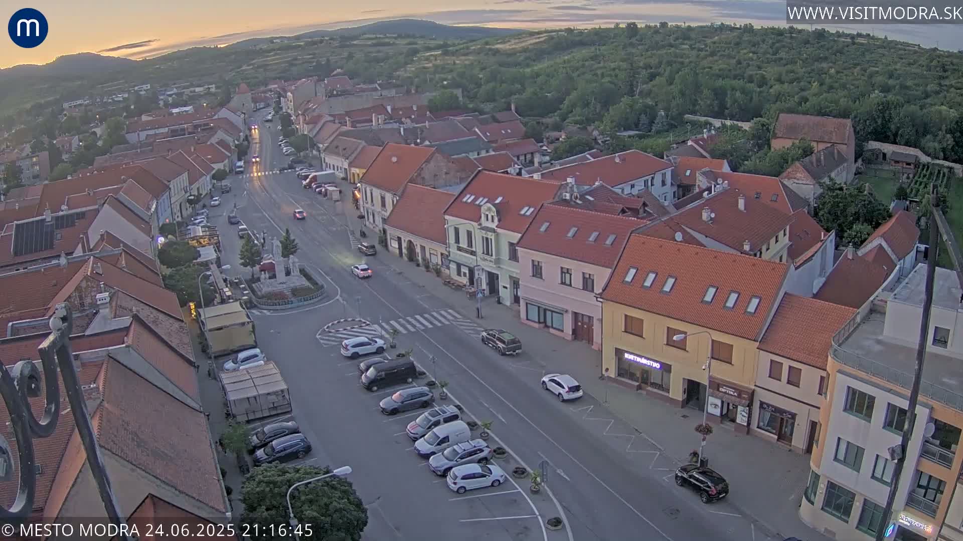 An aerial view of a town square at dusk, showing several parked cars and buildings with red-tiled roofs under a mostly clear sky.