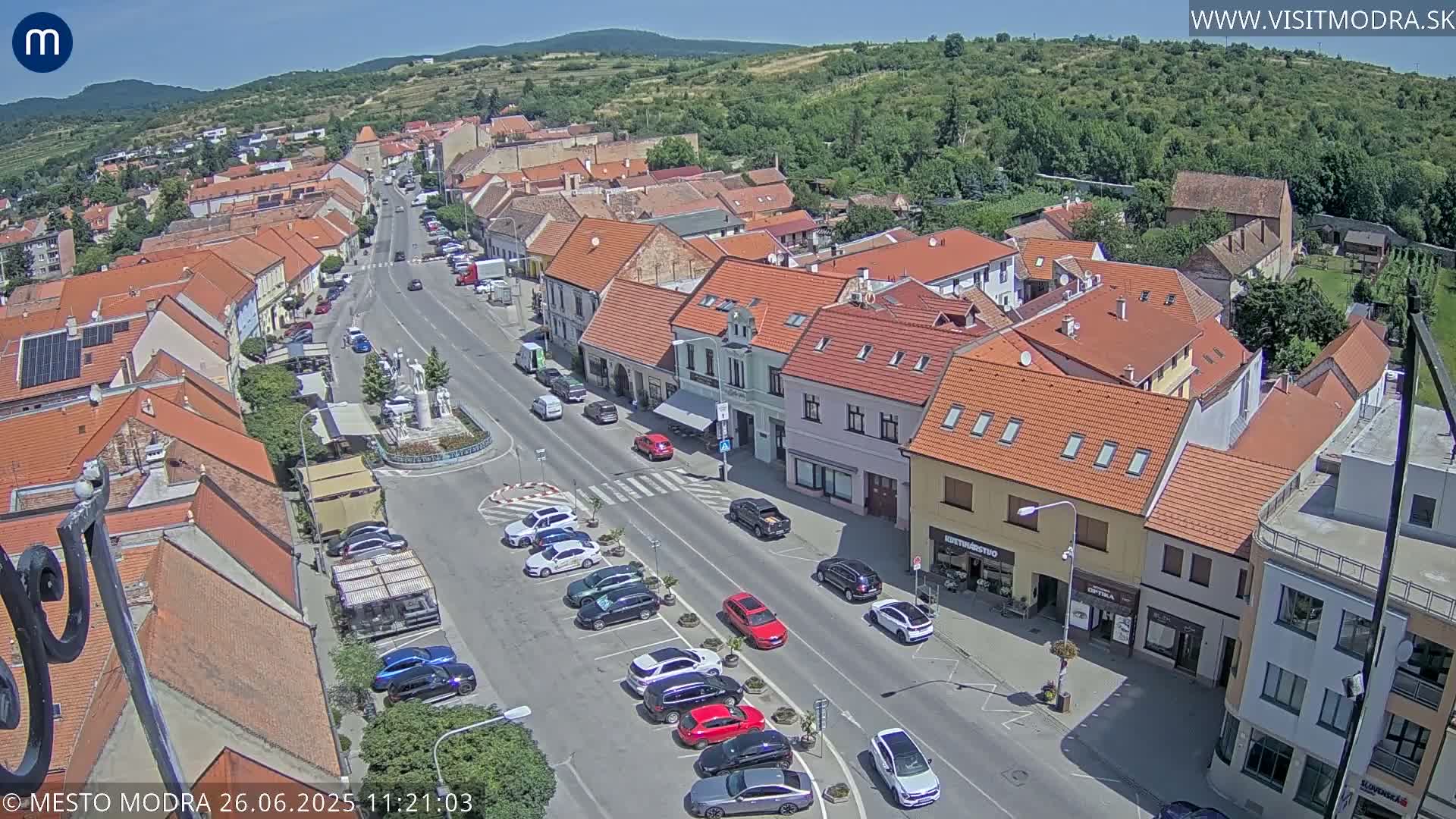 An aerial view of a town square at dusk, showing several parked cars and buildings with red-tiled roofs under a mostly clear sky.