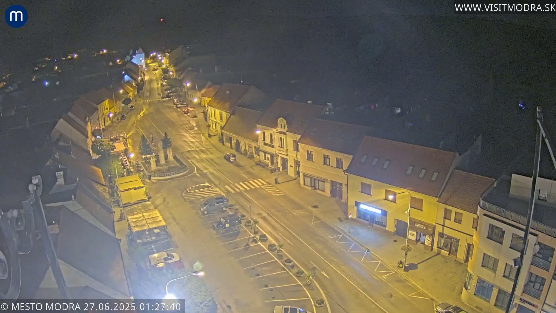 An aerial view of a town square at dusk, showing several parked cars and buildings with red-tiled roofs under a mostly clear sky.