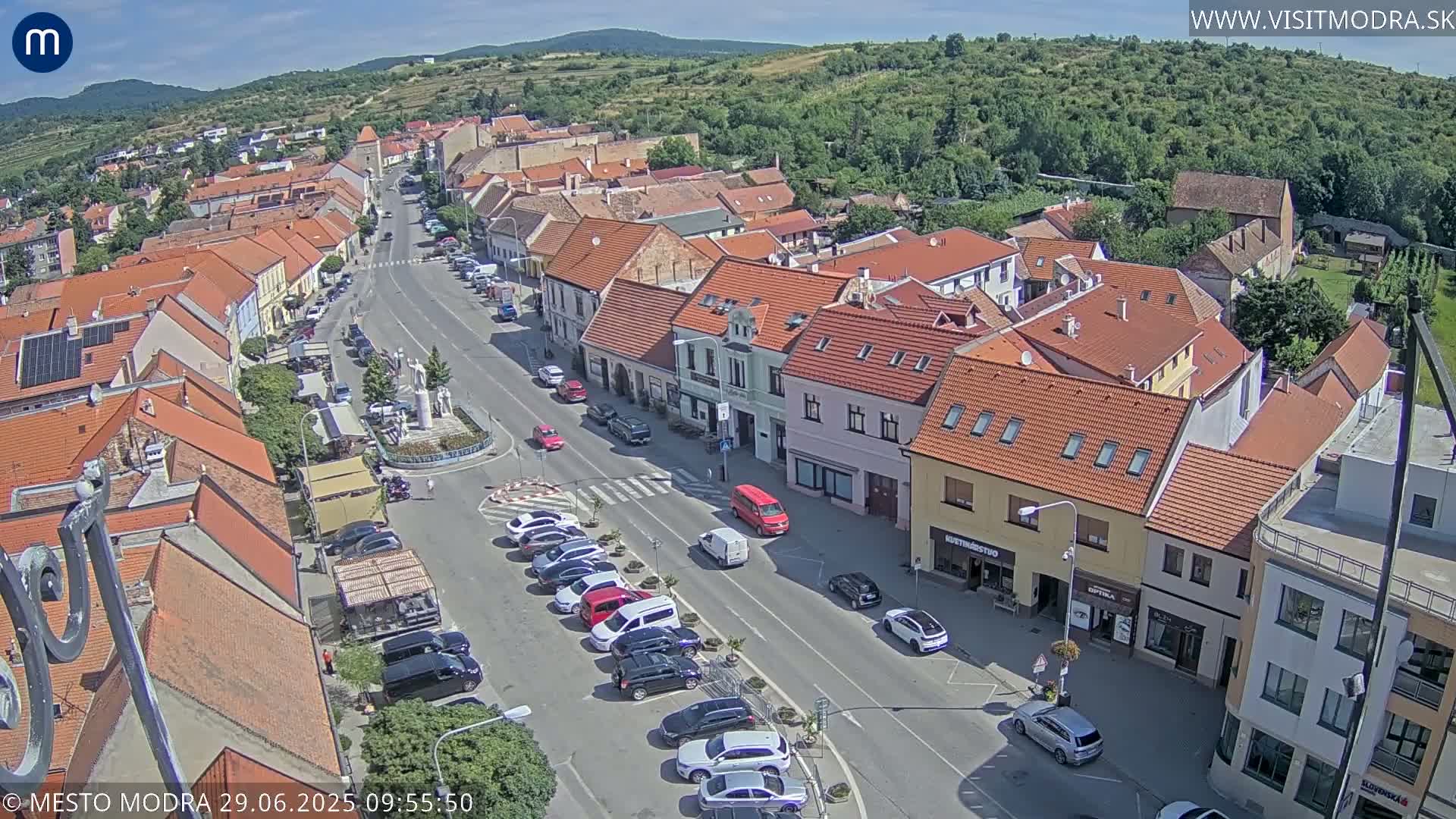 An aerial view shows a town square with cars parked along the sides of a street, surrounded by buildings with red tile roofs under a sunny sky.
