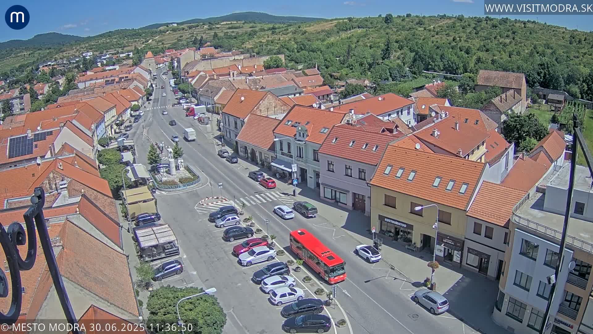 An aerial view of a town square on a sunny day shows a main street lined with parked cars and buildings, with a red bus stopped near the center.