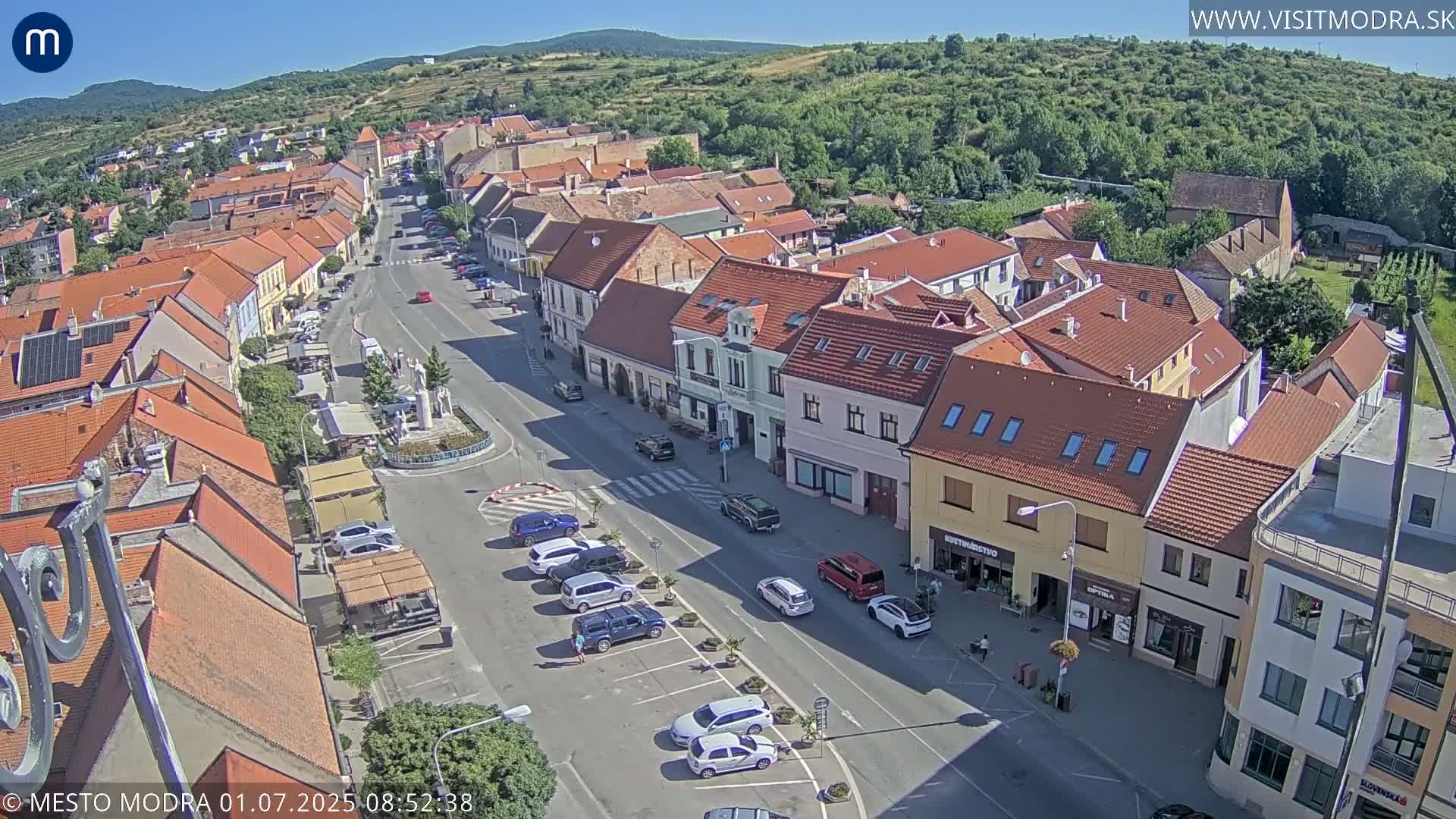 A sunny aerial view of a town's main street lined with buildings, parked cars, and a central square.