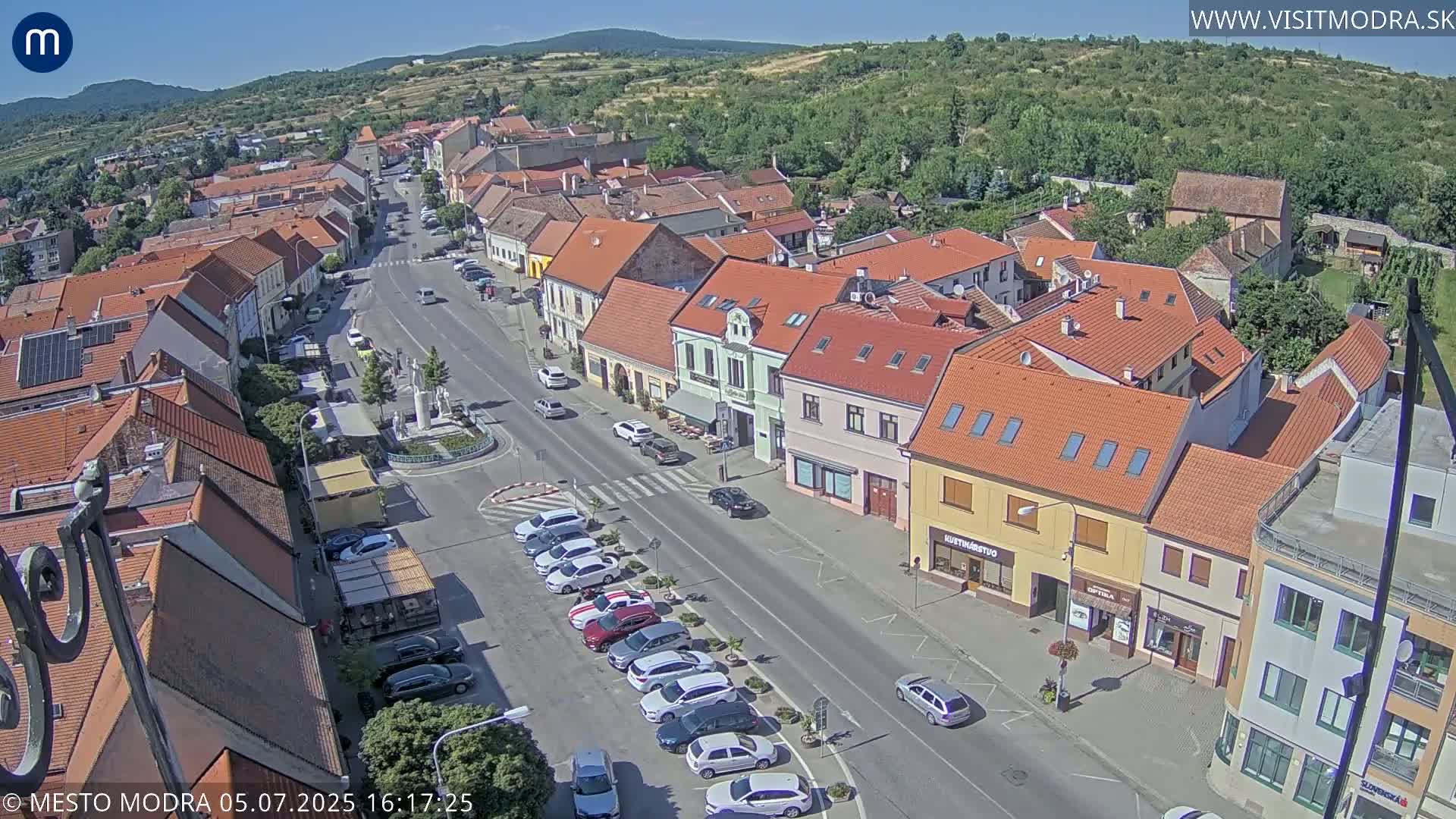 A sunny aerial view shows a town square with parked cars lining a street, surrounded by low, multi-colored buildings and green hills in the distance.