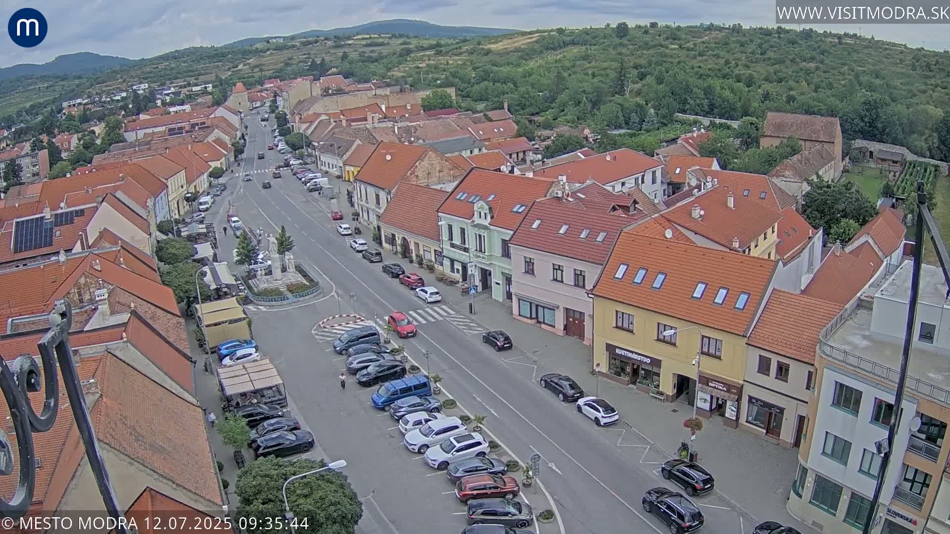 An aerial view shows a town square with parked cars and buildings under overcast skies.