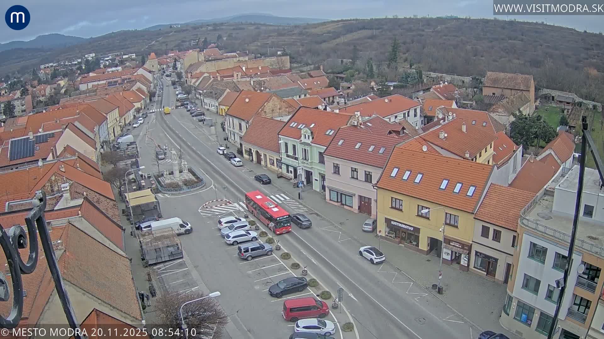 An overcast day blankets a bustling European town street, observed from above, featuring traditional buildings with red-tiled roofs, active vehicle traffic including a red bus, a central monument, and wooded hills in the background.
