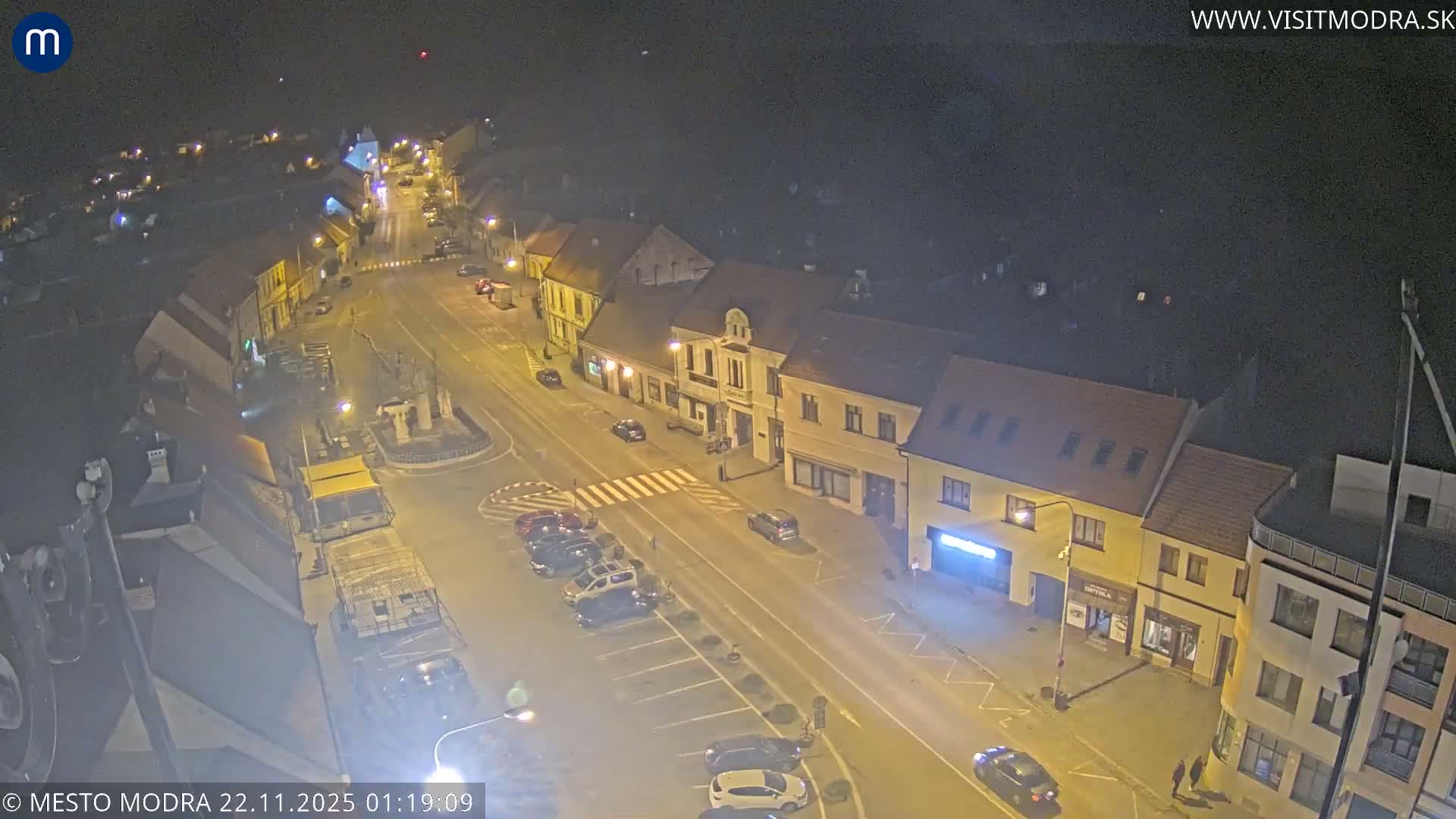 An elevated night view shows a well-lit town street with buildings, parked cars, and a few moving vehicles under a clear, dark sky.