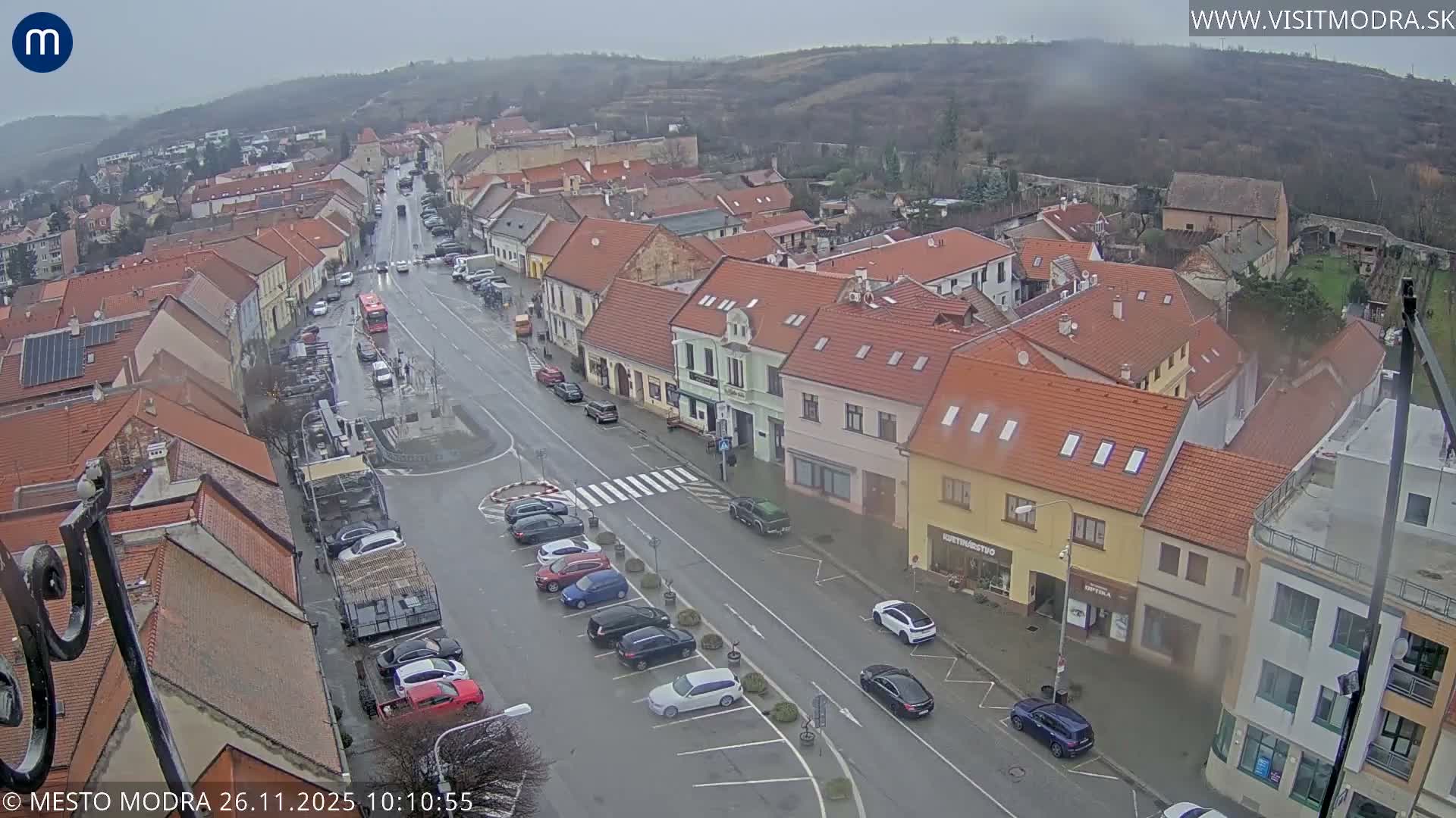An elevated view captures a European town's main street lined with buildings featuring red-tiled roofs, showing active traffic and parked cars on a wet, overcast day with hazy hills in the distance.