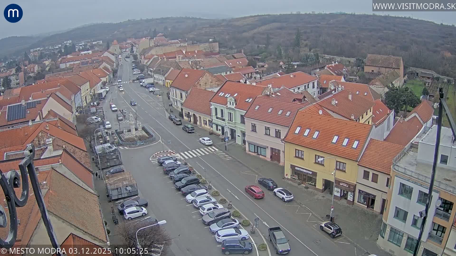 An aerial view captures a bustling European town street lined with buildings featuring predominantly red-tiled roofs, cars parked and driving, and a central monument, all under an overcast sky with hills in the background.