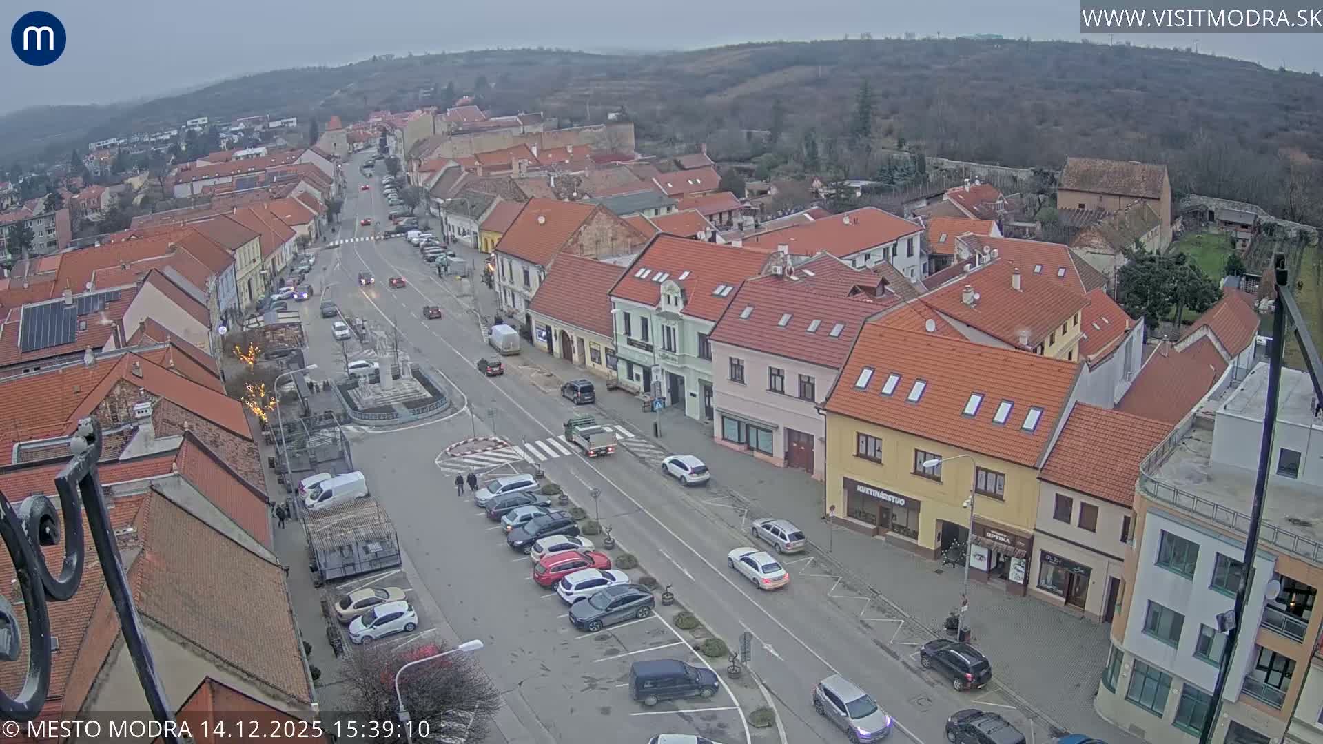 An aerial view captures a bustling European town street lined with buildings featuring predominantly red-tiled roofs, cars parked and driving, and a central monument, all under an overcast sky with hills in the background.