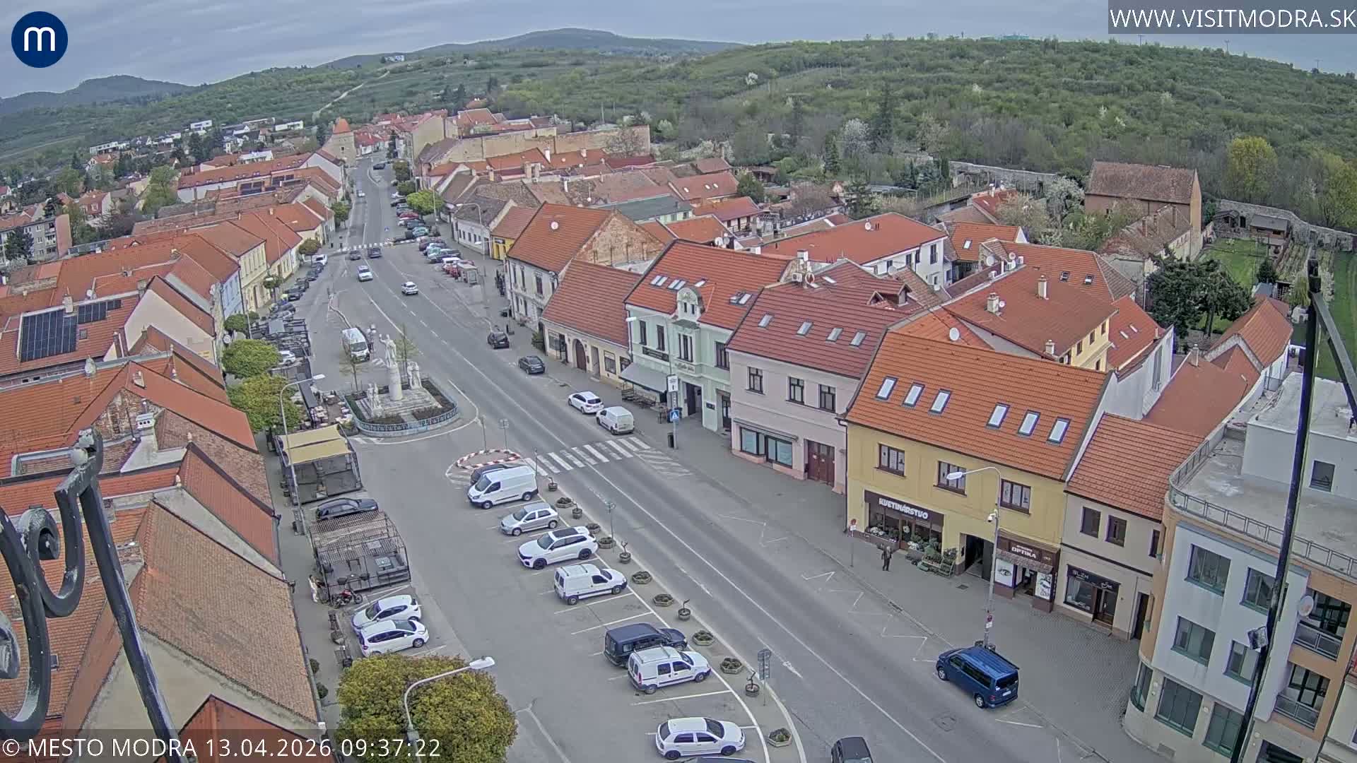 An aerial view captures a bustling European town street lined with buildings featuring predominantly red-tiled roofs, cars parked and driving, and a central monument, all under an overcast sky with hills in the background.
