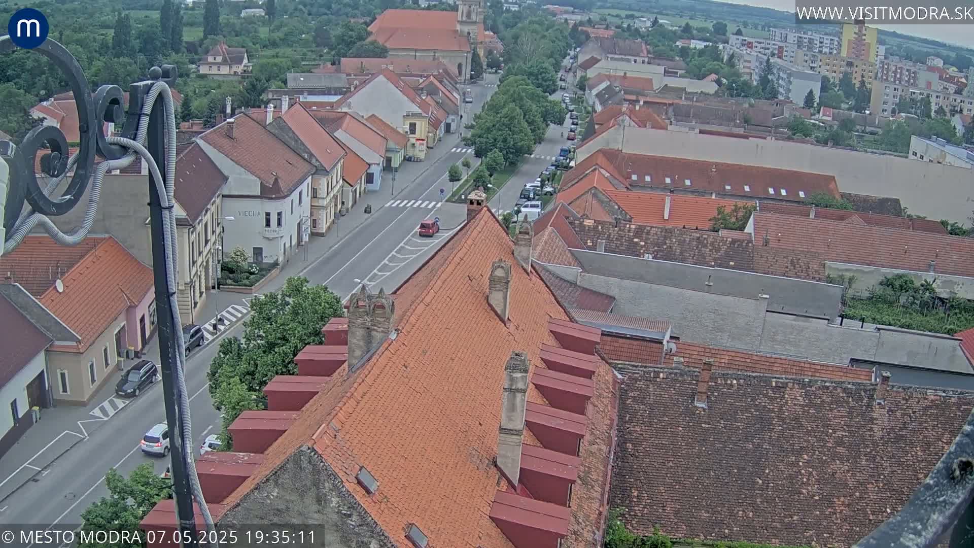 A high-angle, long shot of a town with red-tiled roofs under an overcast sky, showing a street with parked and moving cars.
