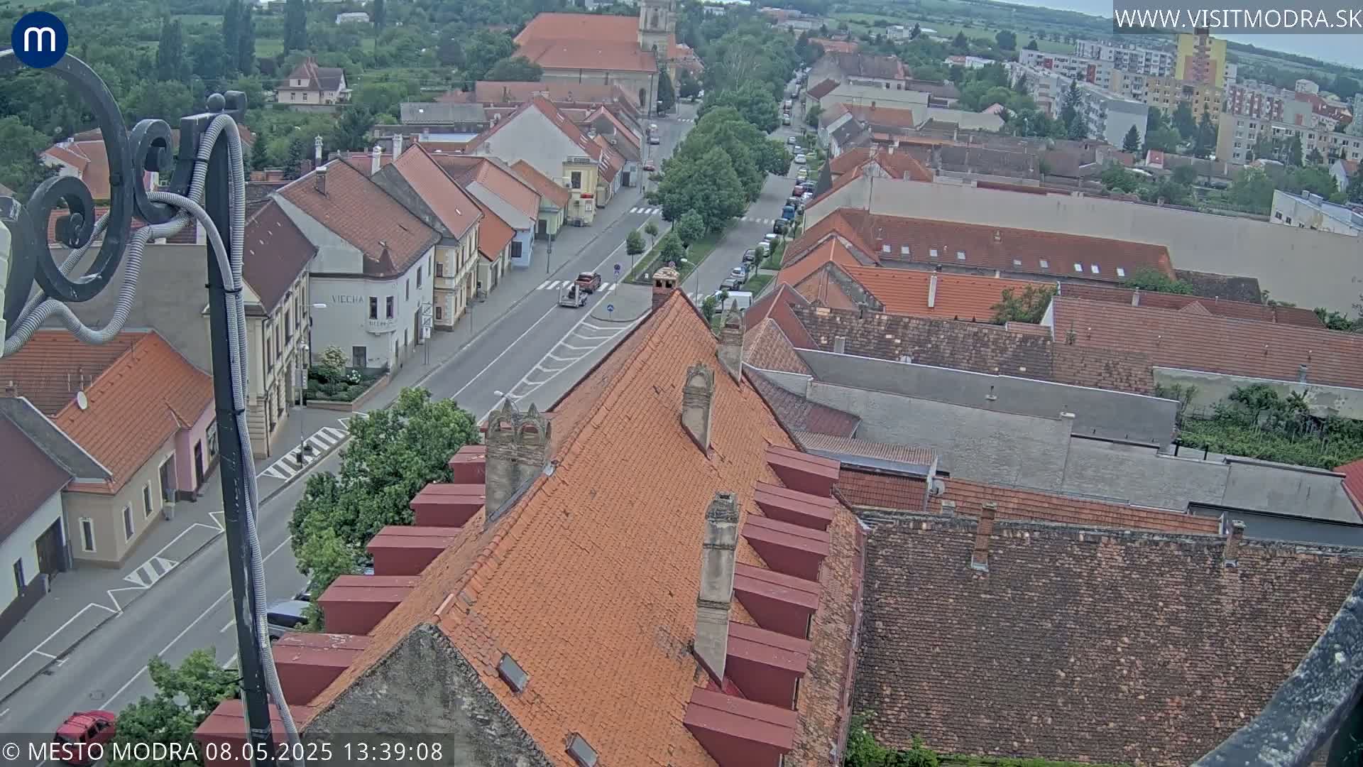 An elevated view of a town on an overcast day shows a street lined with buildings, many with red tile roofs, stretching towards a church in the distance.