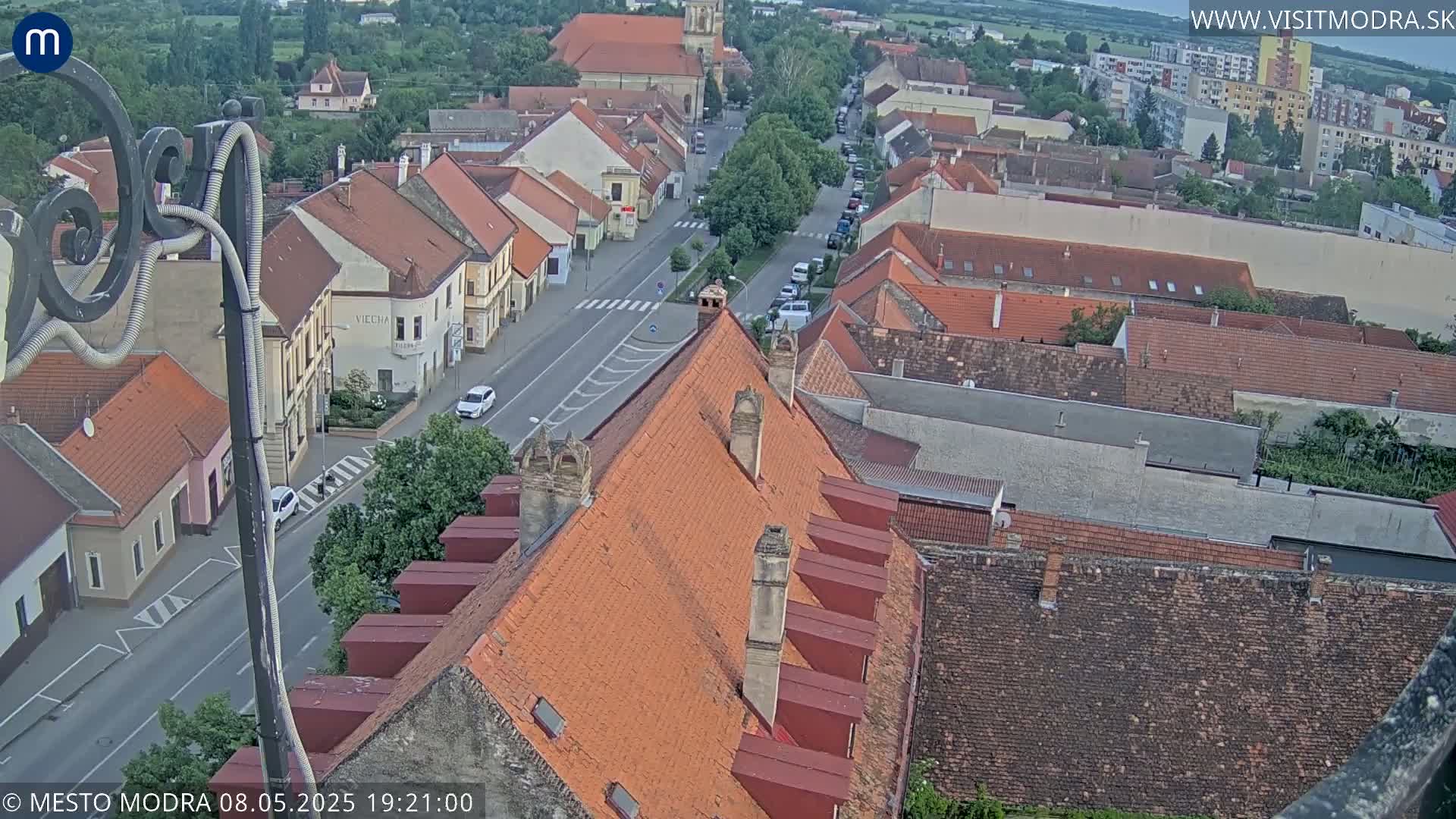 An aerial view shows a town with red-tiled roofs and a street below under a mostly clear evening sky.