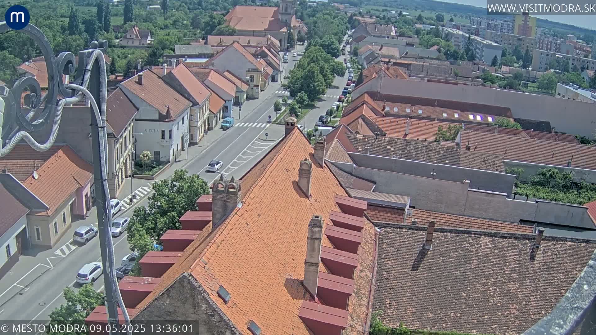 An aerial view of a town on a sunny day, showing numerous buildings with orange-tiled roofs and a street with cars.