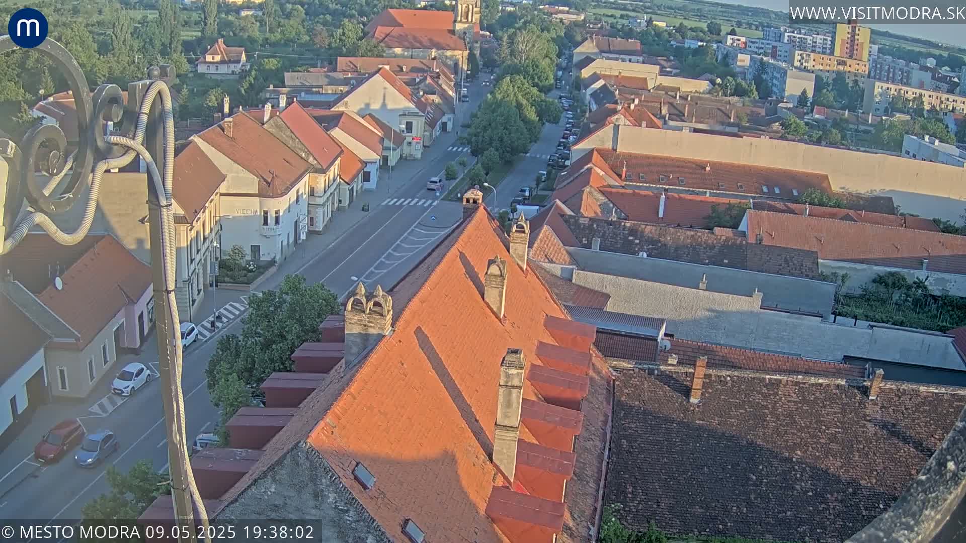 An aerial view of a town shows numerous red-tiled roofs under a partly sunny sky.