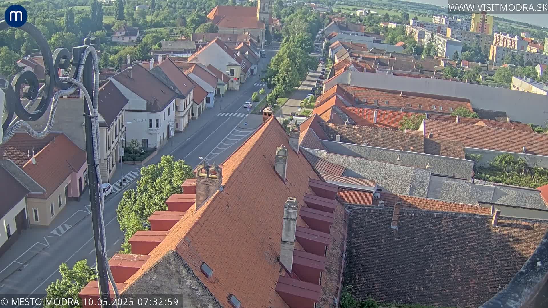 An aerial view of a town on a sunny day shows a street lined with buildings, most with red-tiled roofs, and some taller buildings in the background.