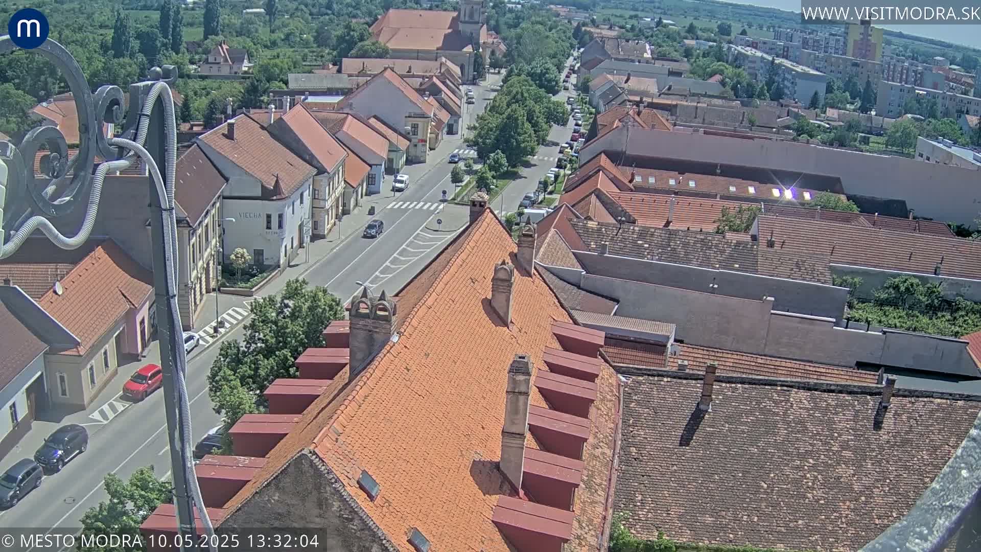 A sunny aerial view shows a town's red-tiled rooftops and a street with cars, surrounded by greenery.