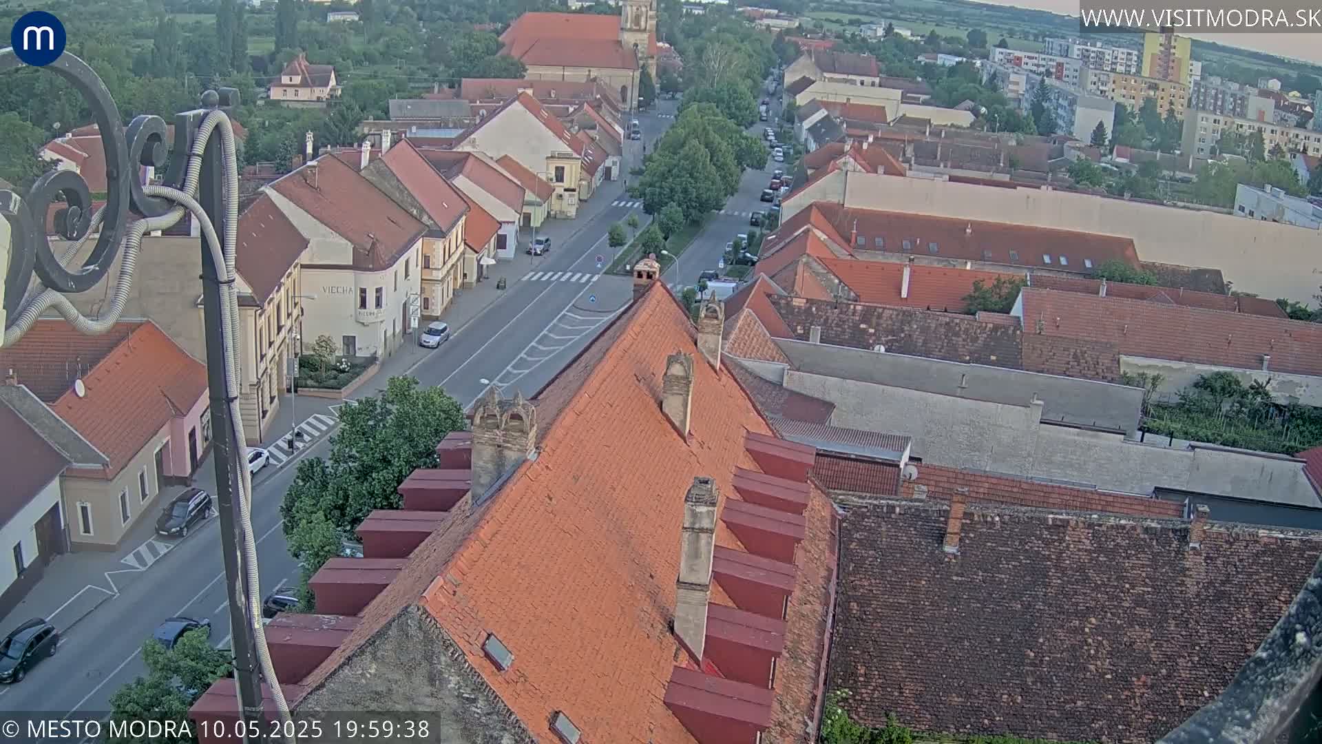 An aerial view of a town's red-tiled rooftops and a street with cars, under a partly cloudy evening sky.