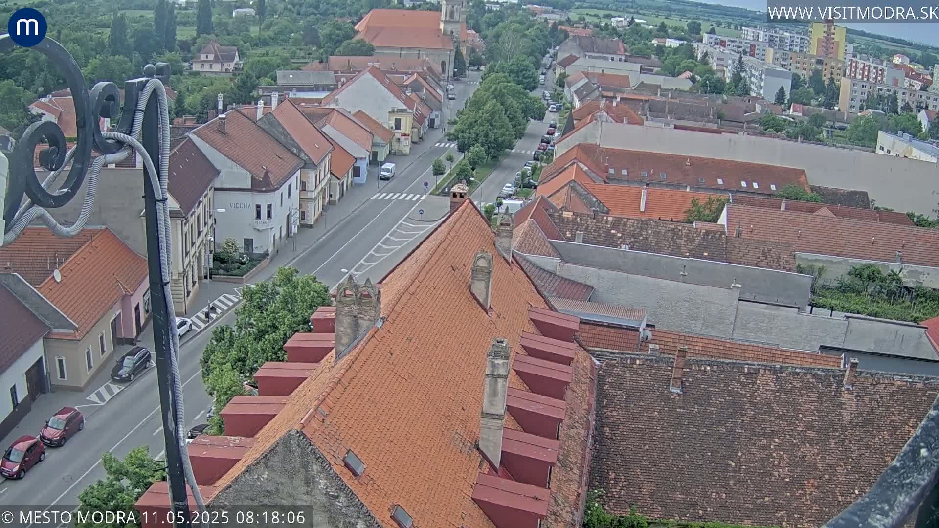 An overcast day reveals a high-angle view of a town's red-tiled rooftops and a street lined with buildings, with cars visible on the road.