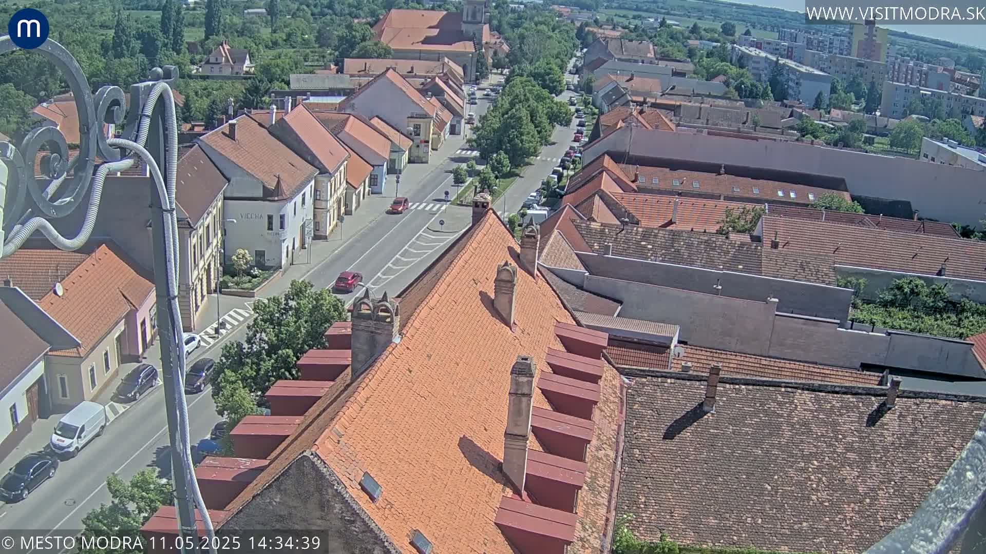 An elevated view shows a town's red-tiled roofs and a street with cars on a sunny day.