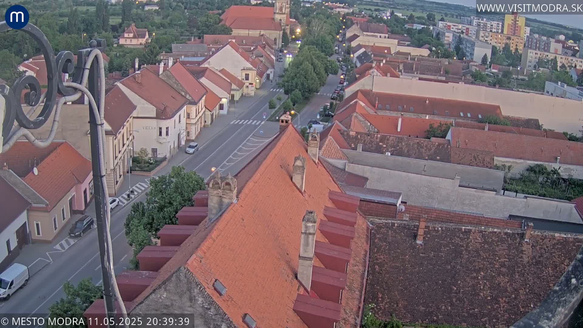An elevated view shows a town's red-tiled rooftops and streets at dusk, under a clear sky.