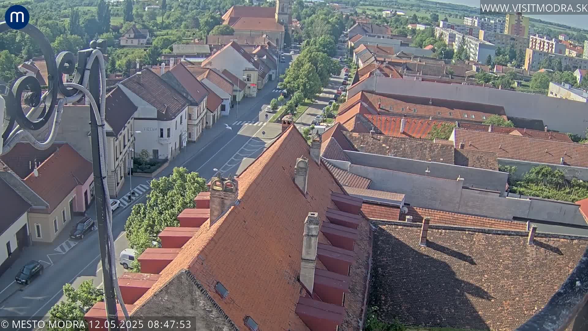 A high-angle view of a town on a sunny day, showing red-tiled rooftops, a street with cars, and surrounding greenery.