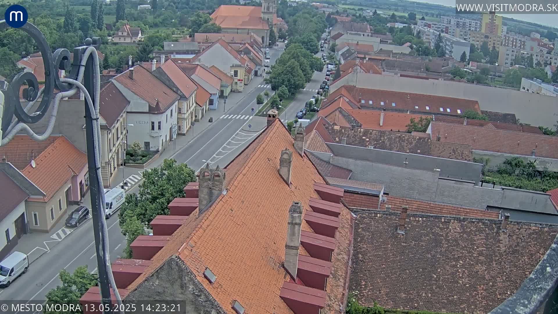 An elevated view of a town on a partly cloudy day, showing numerous red-tiled rooftops and a street with parked cars.