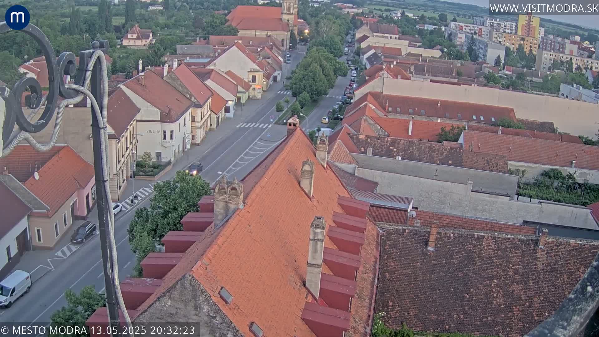 An elevated view shows a town's red-tiled rooftops and streets at dusk, under a clear sky.