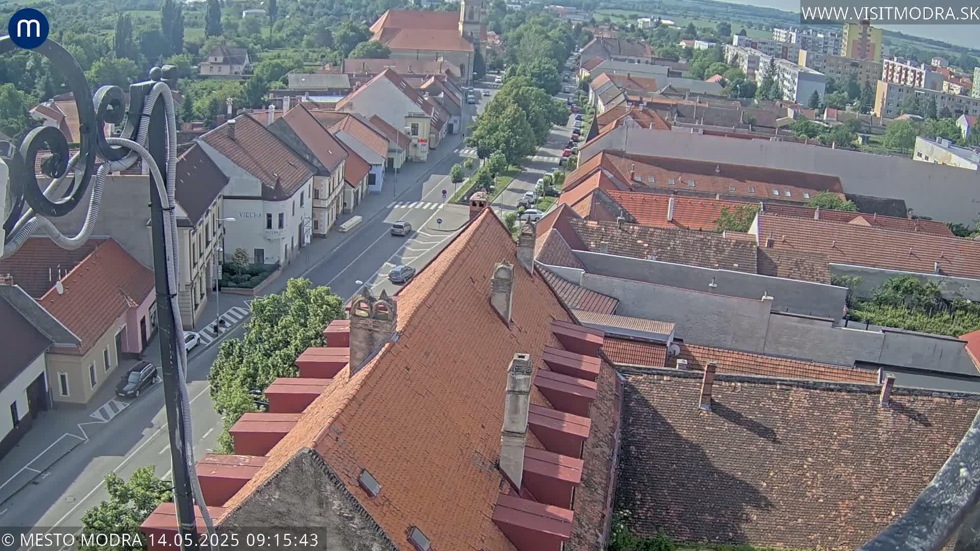 An aerial view shows a town on a sunny day, with red-tiled roofs dominating the cityscape and a few cars visible on the streets below.