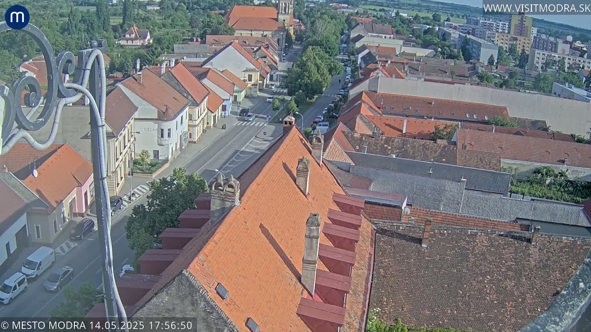 An aerial view shows a town with red-tiled roofs and a street below, on a sunny day.