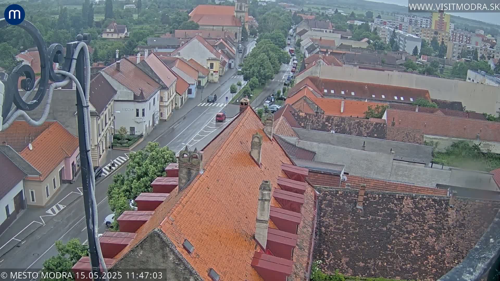 An elevated view of a town on an overcast day shows a street with cars and numerous buildings with red-tiled roofs.