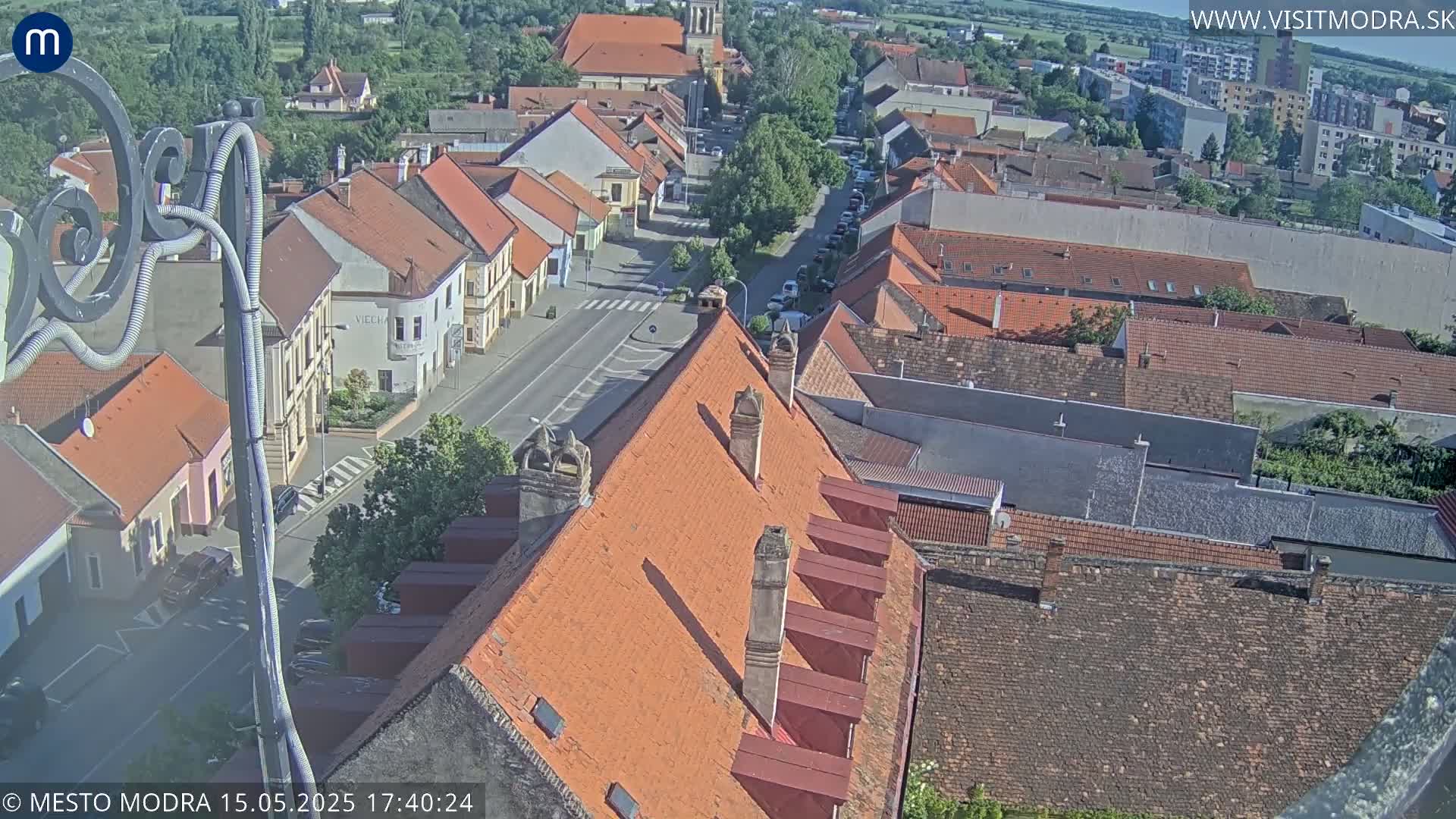 An elevated view of a town on a sunny day shows a collection of red-tiled roofs and a street with cars below.