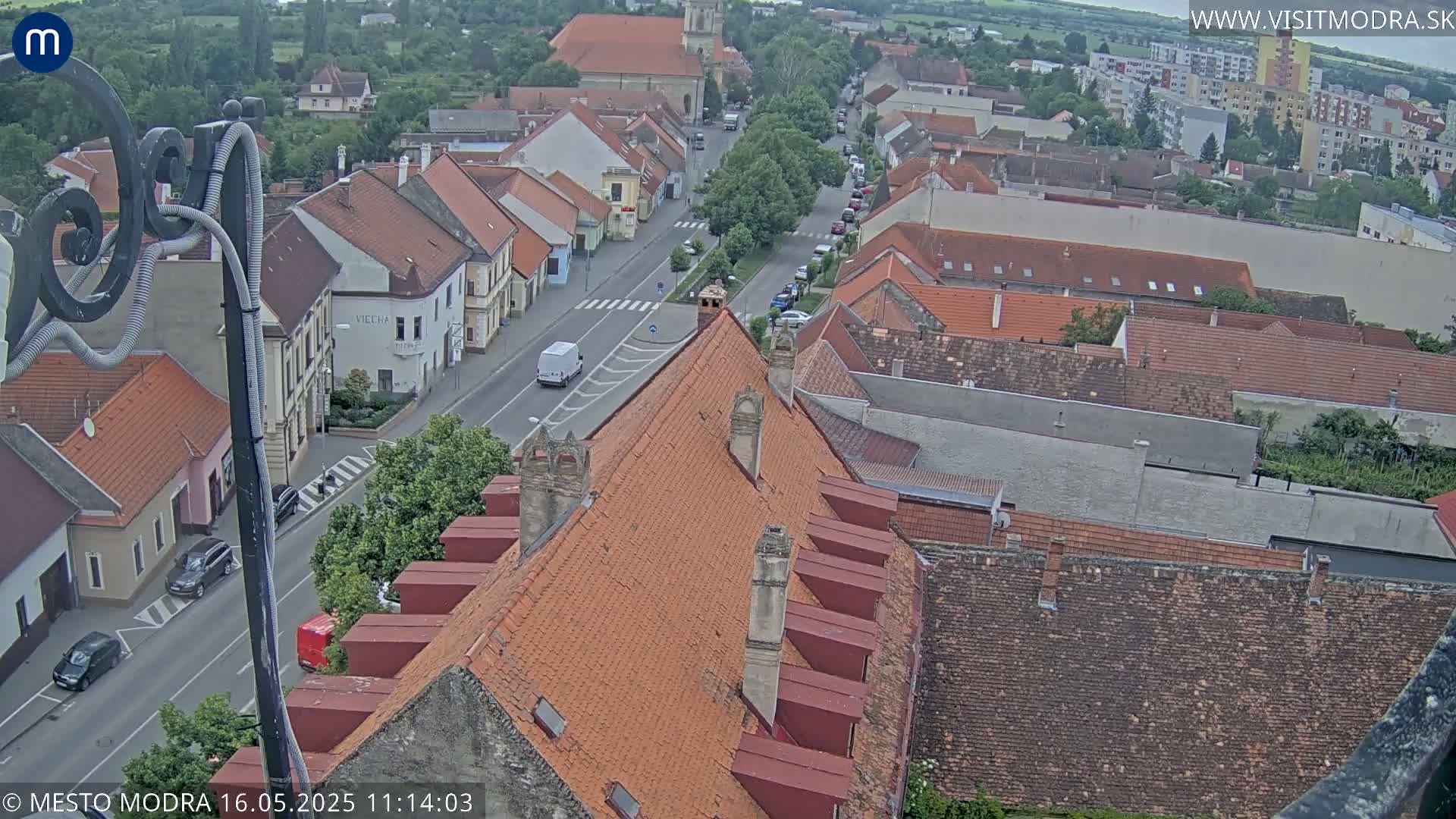 An overcast view of a town's rooftops and a street with cars, showing a mix of building styles and a primarily red-tiled roofscape.
