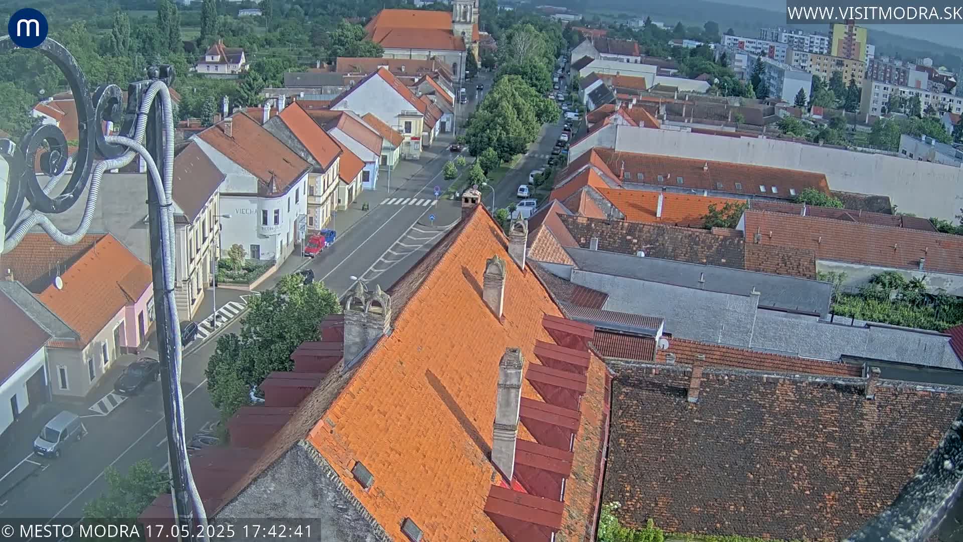 An aerial view of a town's red-tiled rooftops and streets on a partly cloudy day.