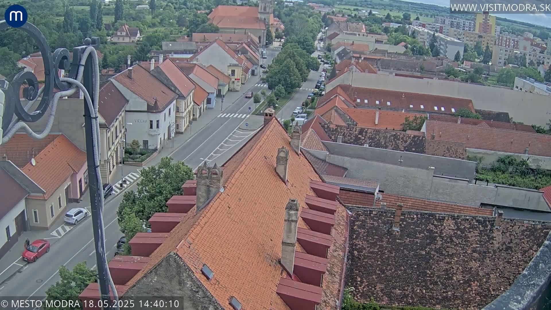 A high-angle view of a town's street and rooftops on a partly cloudy day, showing cars parked along the road and numerous red-tiled roofs.