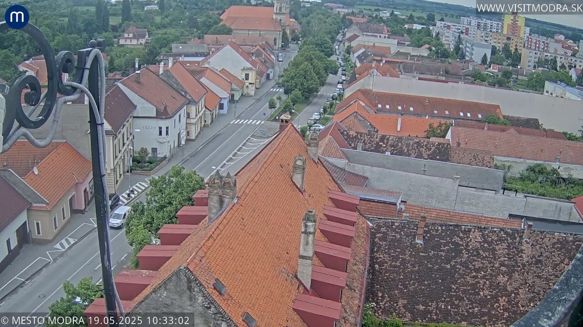 An overcast day reveals a high-angle view of a town's red-tiled rooftops and a street lined with buildings, extending to a distant horizon.