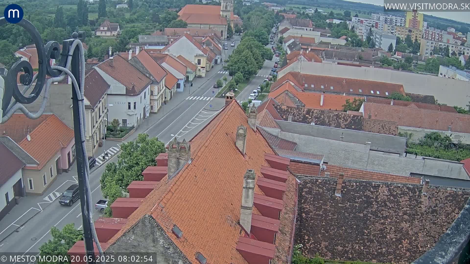 An aerial view shows a town with red-tiled roofs and a street lined with buildings under an overcast sky.