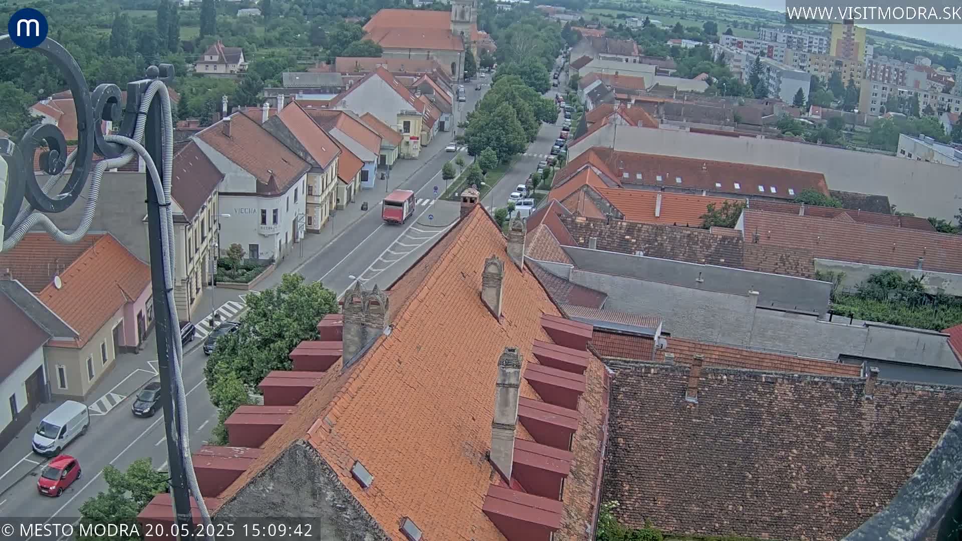 An elevated view of a town's street lined with closely-packed buildings with red-tiled roofs, under a partly cloudy sky.