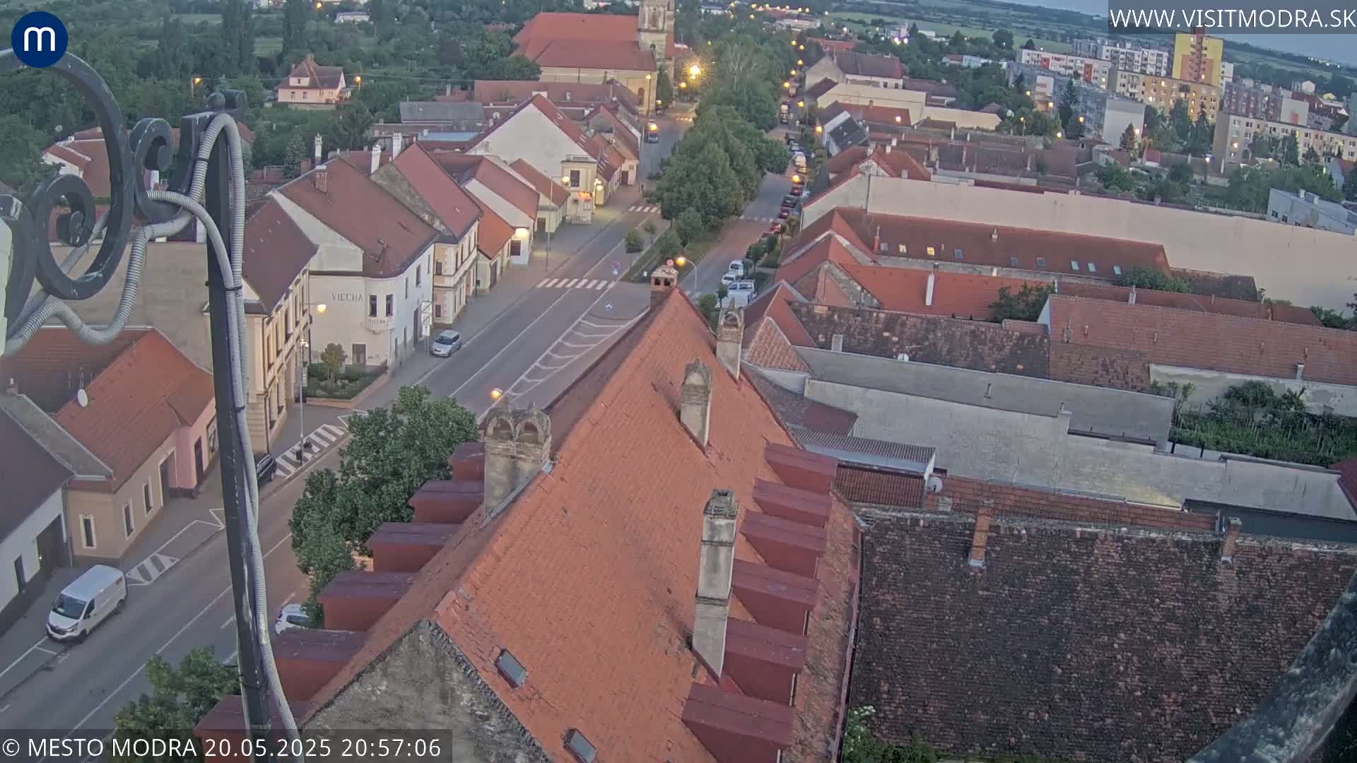 An elevated view of a town at dusk, showing a street with cars and many closely packed buildings with reddish-brown roofs.