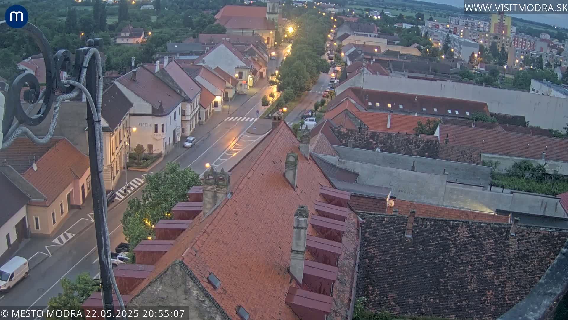 An elevated view of a town at dusk, showing a street with cars and buildings with red tile roofs under a twilight sky.