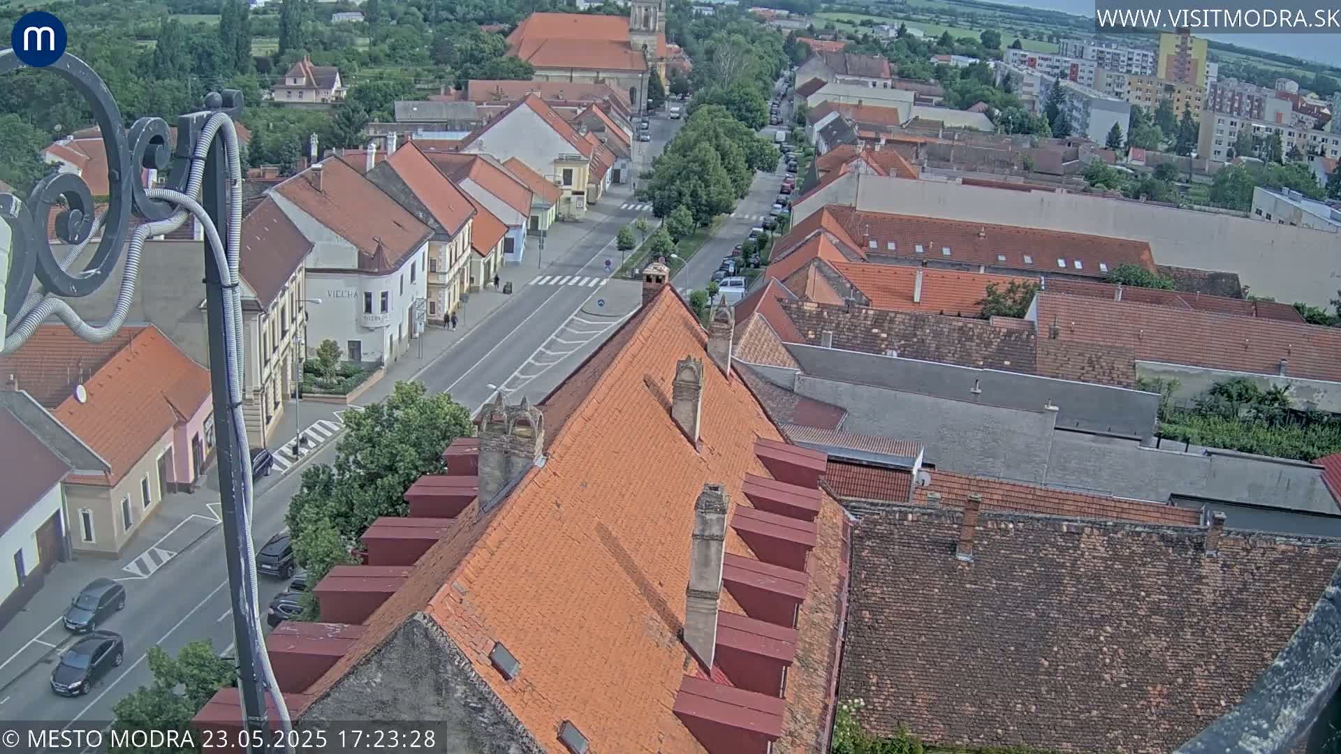 An overcast day reveals a high-angle view of a town's red-tiled rooftops, streets, and buildings, with cars parked along the roads.