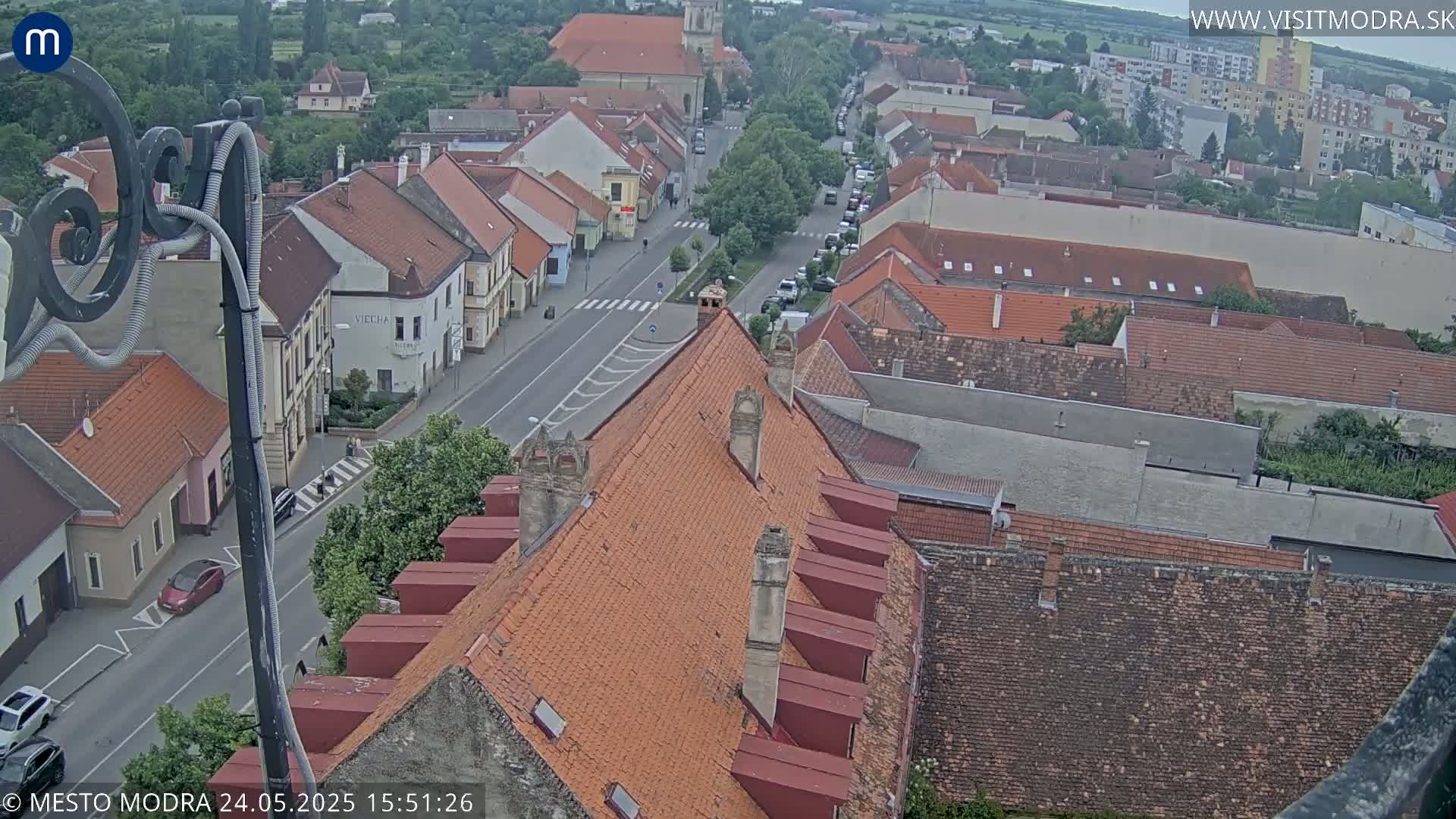 An overcast day reveals a high-angle view of a town's streets and red-tiled rooftops, with cars parked along the road.