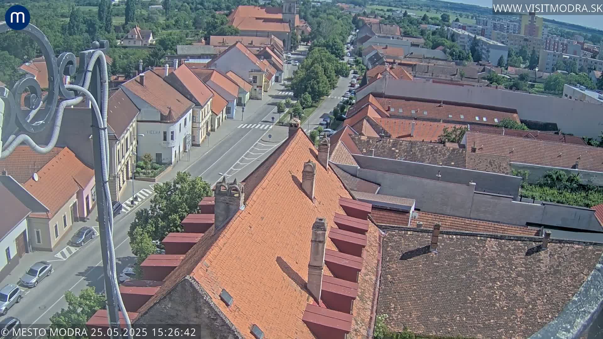 An elevated view of a town on a sunny day shows a street lined with buildings and many red-tiled roofs.