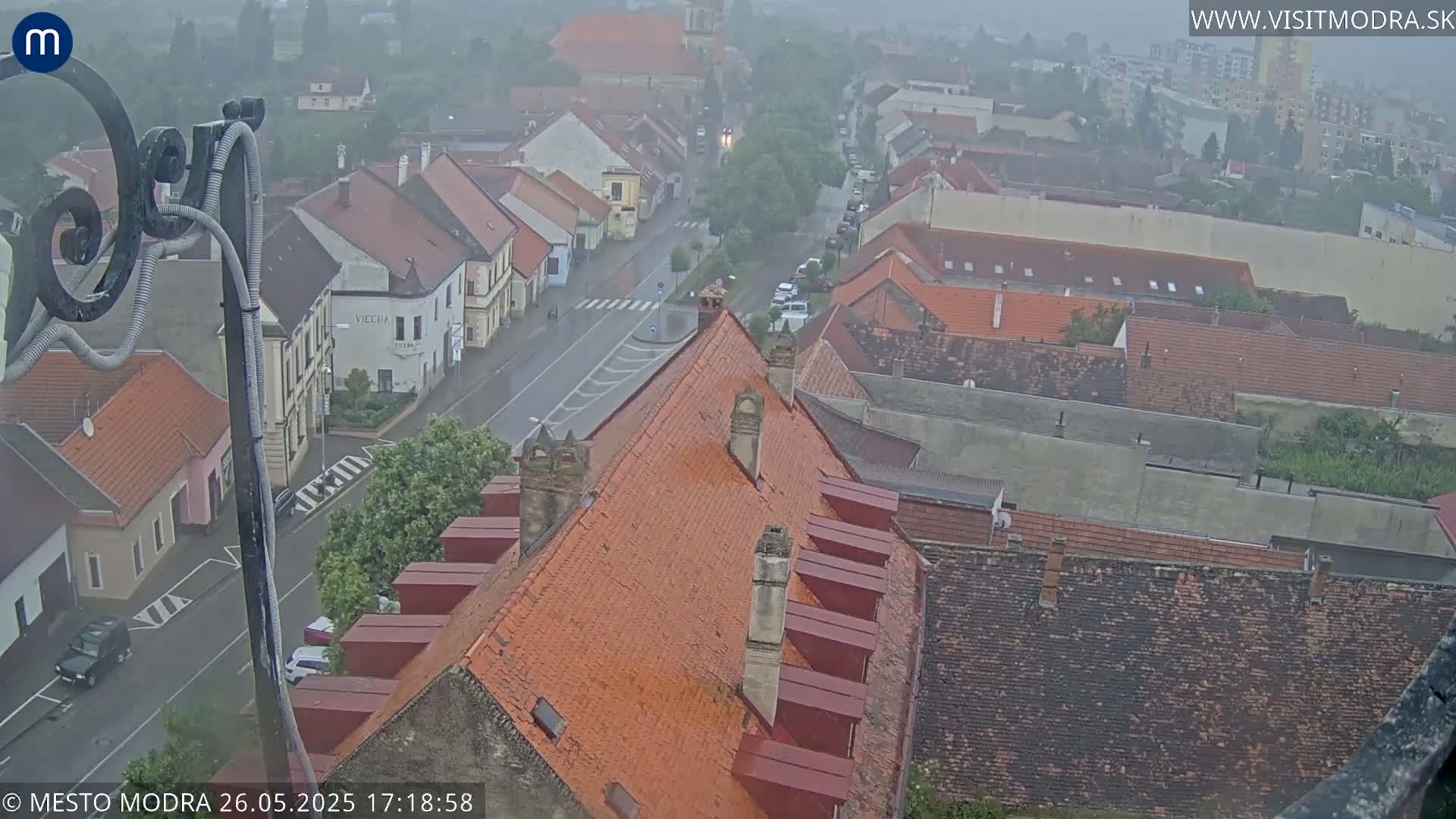 A high-angle view of a town on a rainy day, showing red-tiled roofs, a street with sparse traffic, and a hazy atmosphere.